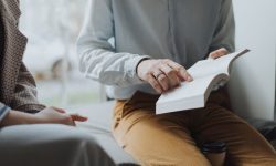 Mann in weißem Hemd und brauner Hose sitzt auf einem weißen Stuhl und liest ein Buch