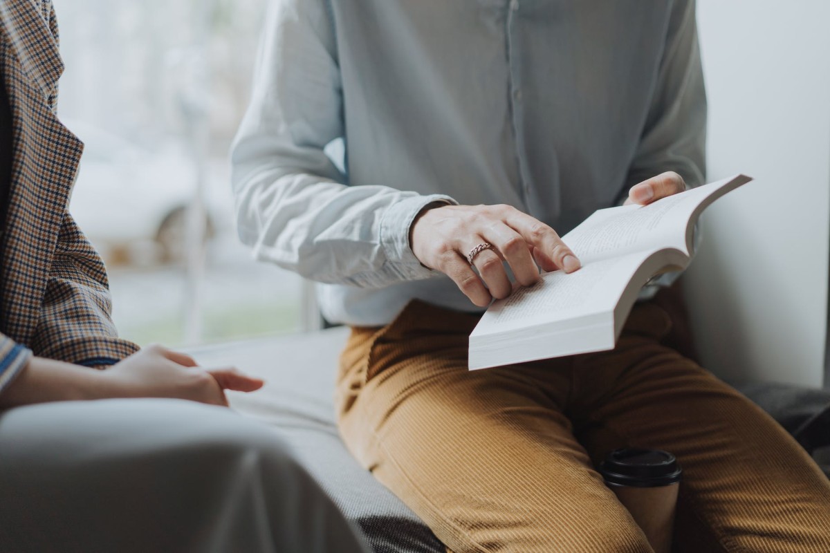hombre con camisa de vestir blanca y pantalones marrones sentado en una silla blanca leyendo un libro