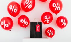 um saco de papel preto com etiqueta de venda no meio de balões vermelhos com símbolos de porcentagem em fundo branco