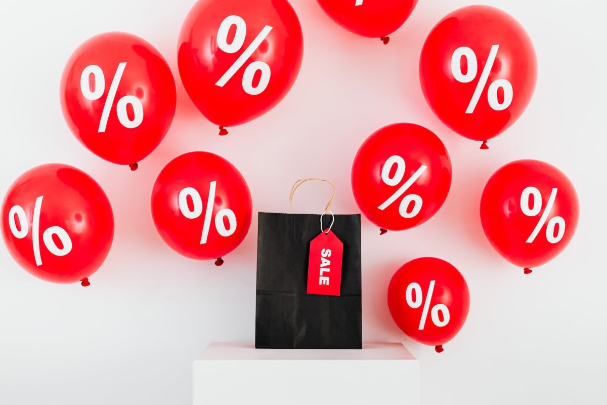 una bolsa de papel negra con etiqueta de venta en medio de globos rojos con símbolos de porcentaje sobre fondo blanco